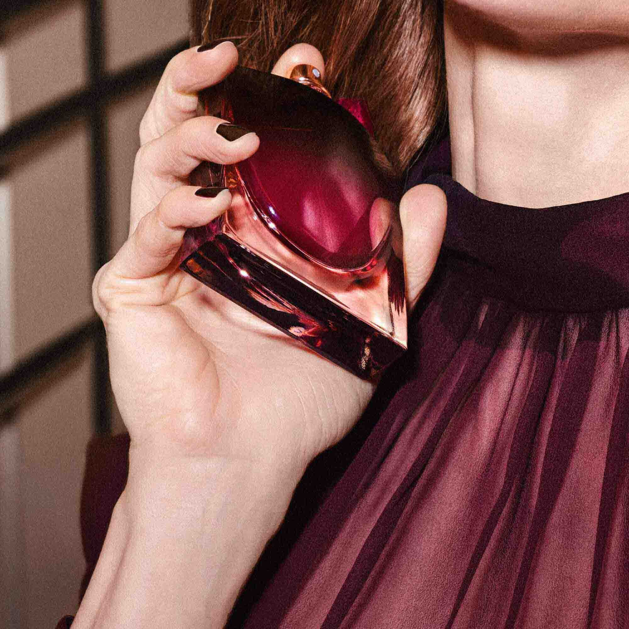 A person with dark nail polish holds the dark red perfume bottle gracefully against their neck while wearing a burgundy pleated garment.	