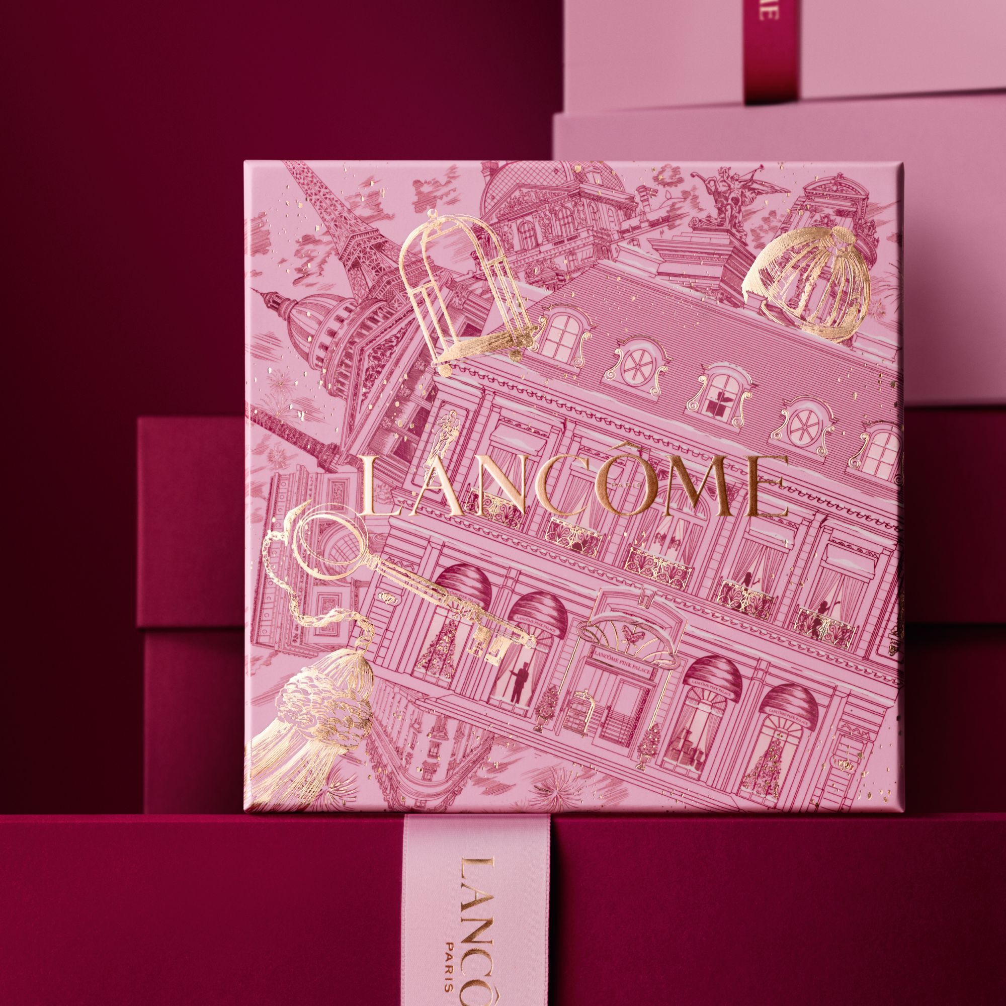 Image of pink Lancome holiday gift box standing in front of a pile of pink and dark red gift boxes, set against a dark red background. 